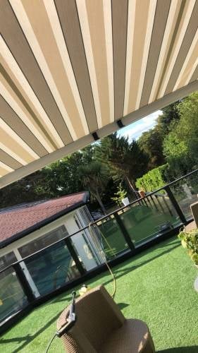 Retracted striped awning over a patio with a green lawn and garden furniture overlooking trees and landscaping