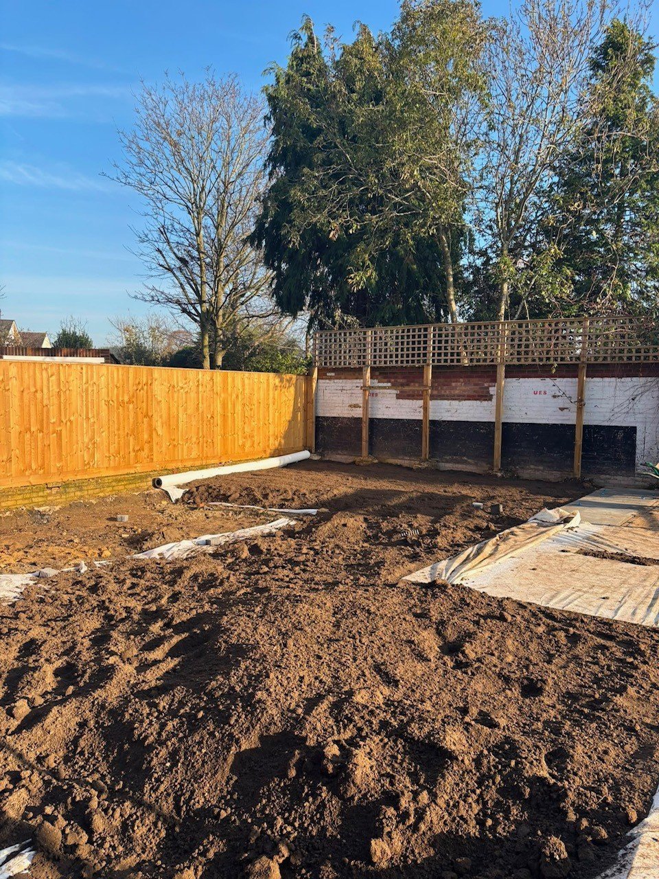 Residential garden under construction with piles of soil, wooden fencing, and bare trees against blue sky
