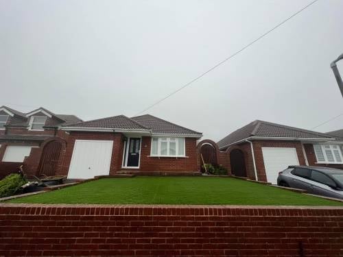 Red brick bungalow with white garage doors, well-maintained green lawn, and neighboring houses on overcast day