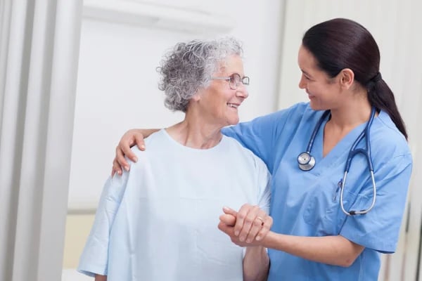 A smiling female healthcare worker in blue scrubs and stethoscope guides an elderly woman in a light blue shirt during a medical consultation.
