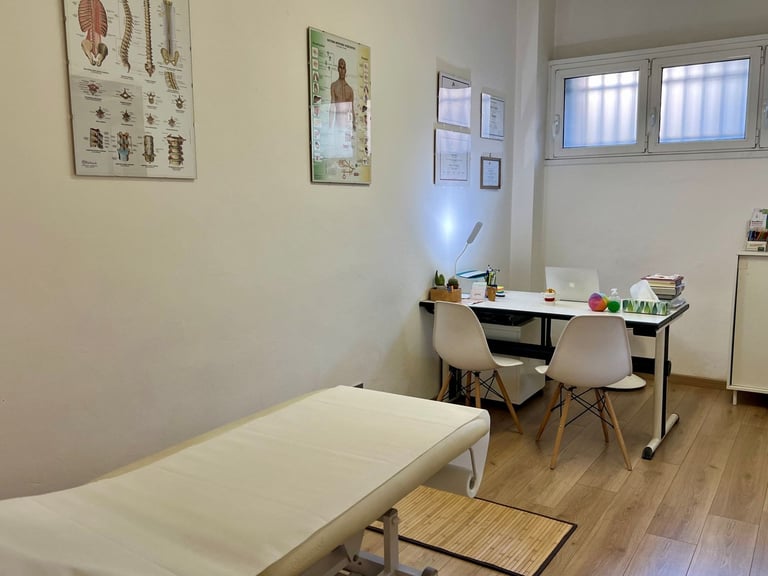 A bright medical examination room with a treatment bed, desk with rolling chair, anatomical posters on walls, and windows with blinds