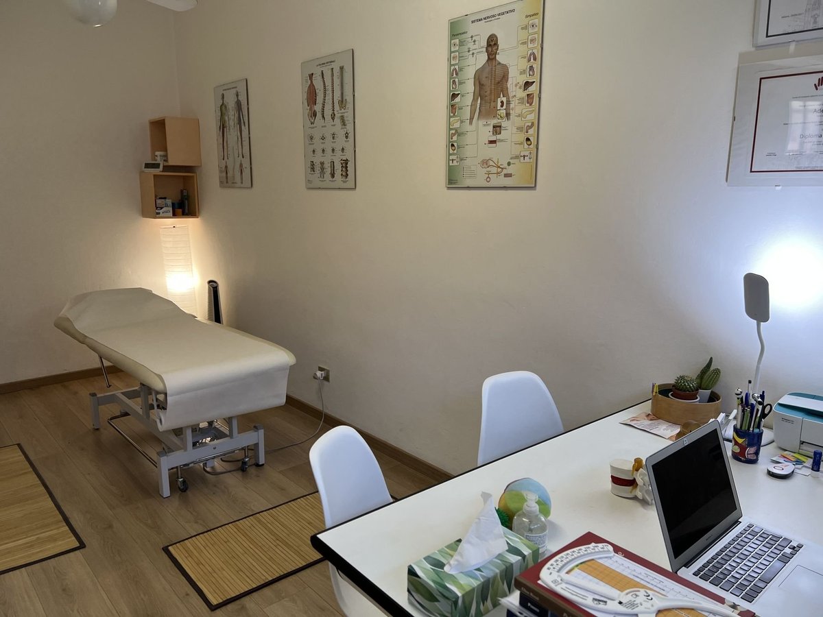 Medical clinic office with treatment bed, desk with laptop and chairs, anatomical charts on walls