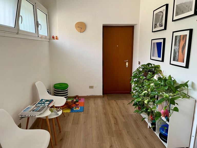 Bright home interior with wooden door, small table with stacked bowls and magazines, potted plant, wall clock, and framed artwork on white walls