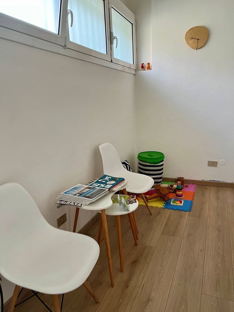 Bright minimalist playroom with two white chairs, small tables with toys, colorful puzzle mat, and wooden clock on wall