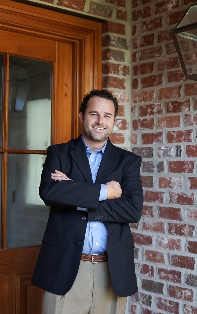 Professional man in navy blazer and light blue shirt posing with arms crossed against red brick wall next to wooden door