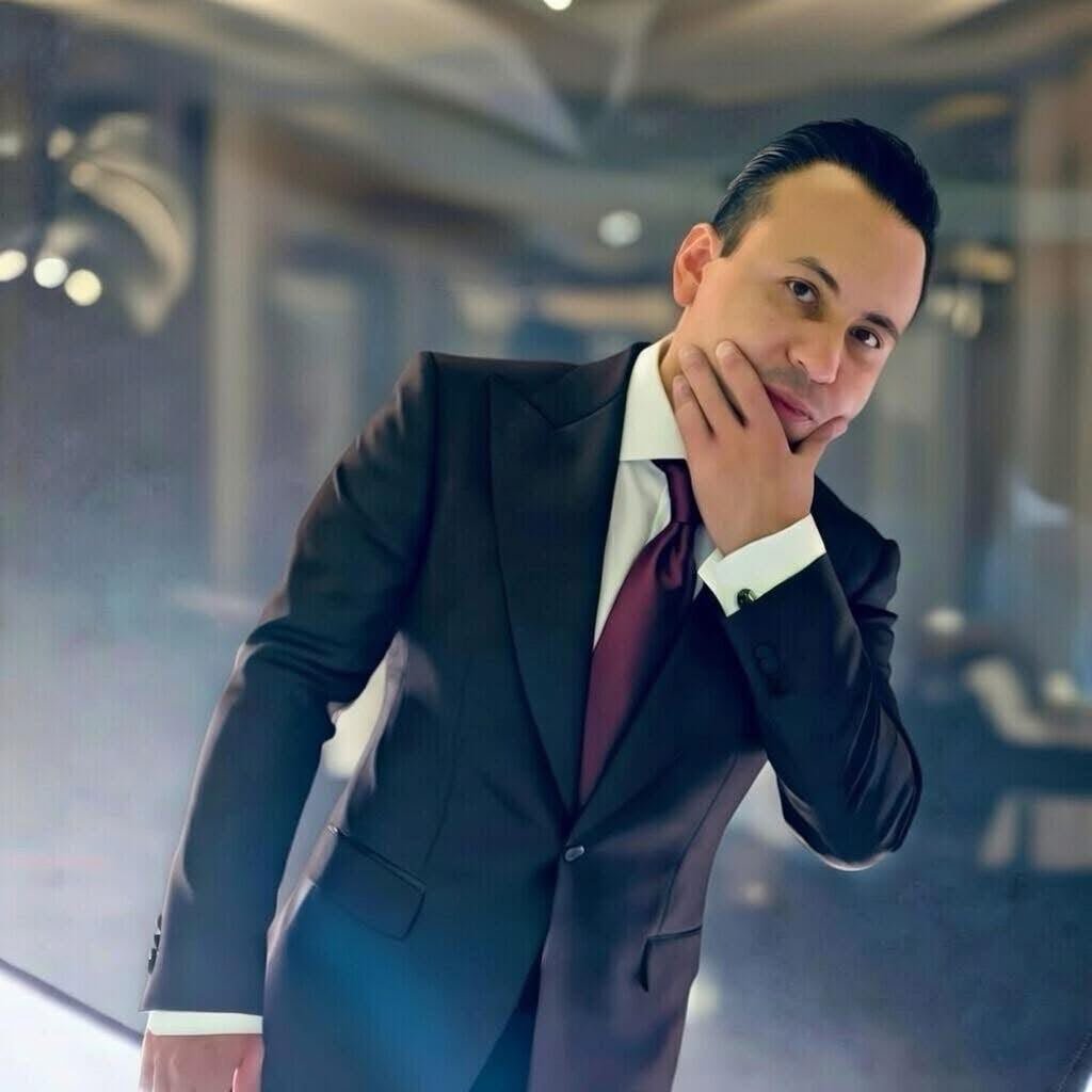 Professional man in dark suit and burgundy tie posing thoughtfully with hand to chin in modern office interior
