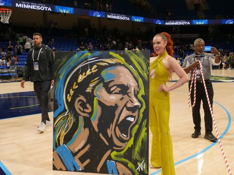 Woman in yellow dress stands next to large portrait artwork on NBA court during game event
