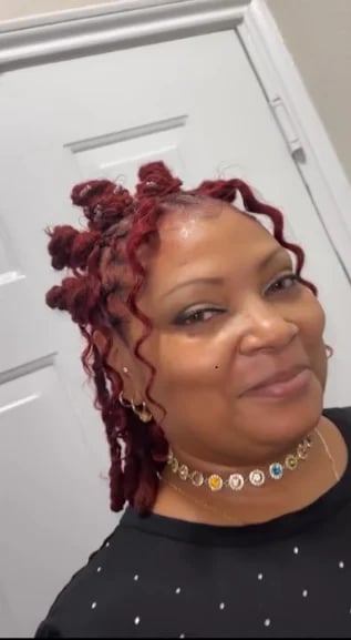 Woman with burgundy curled hair and gold rhinestone necklace wearing a black studded top, smiling at the camera