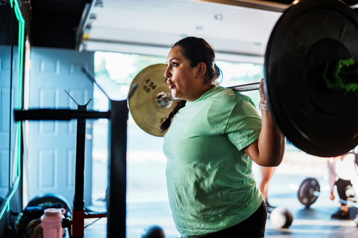 Woman in green shirt performing barbell squat exercise in gym with weights loaded on bar