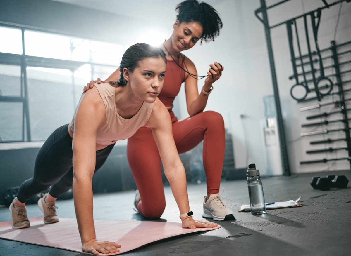 Two women working out together in a gym, one in a plank position and one standing behind her in sportswear