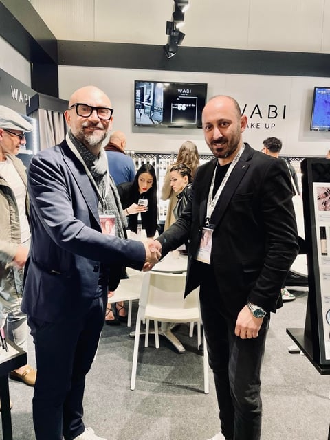 Two men in business suits shaking hands at a WABI trade show booth, wearing Cosmoprof name badges