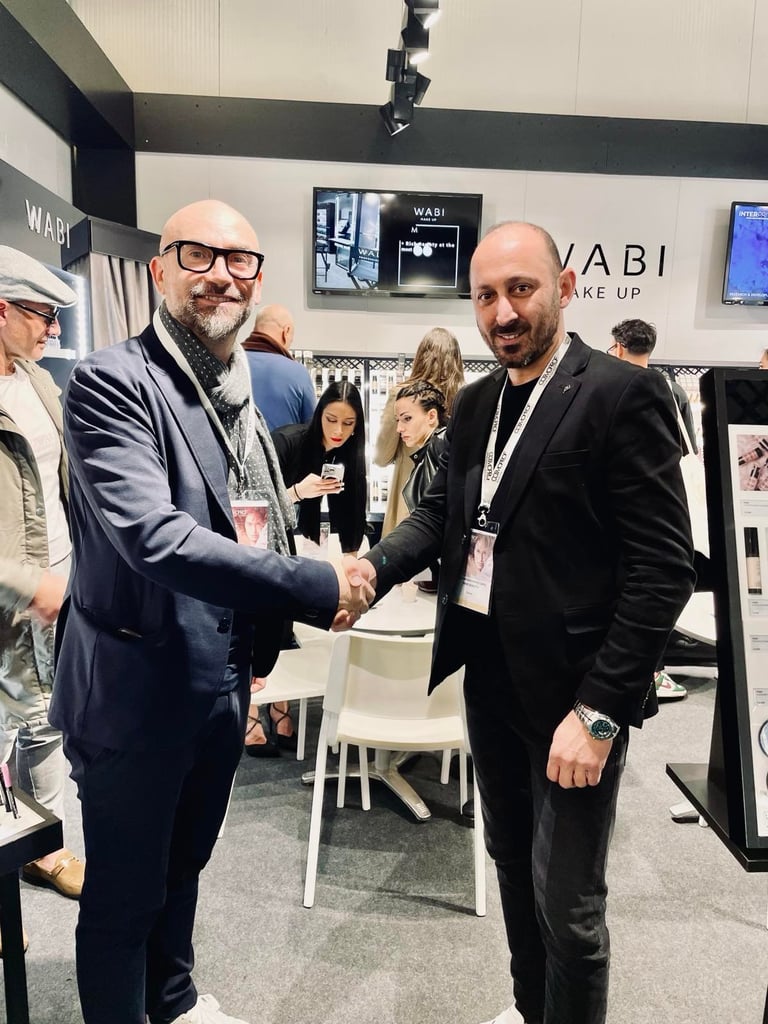 Two men in business suits shaking hands at a WABI trade show booth, wearing Cosmoprof name badges