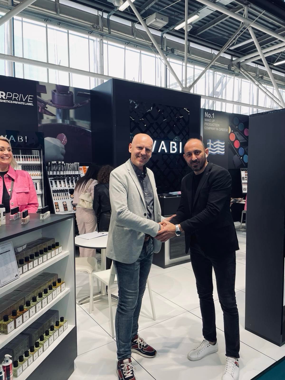 Two men shake hands at a retail cosmetics booth labeled ABI at a trade show or exhibition