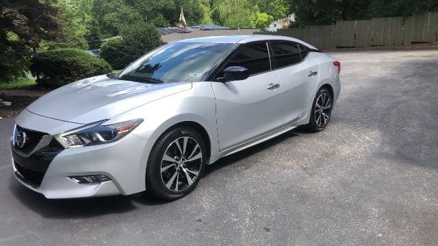 White Nissan Maxima parked on an asphalt driveway with trees and fence in the background