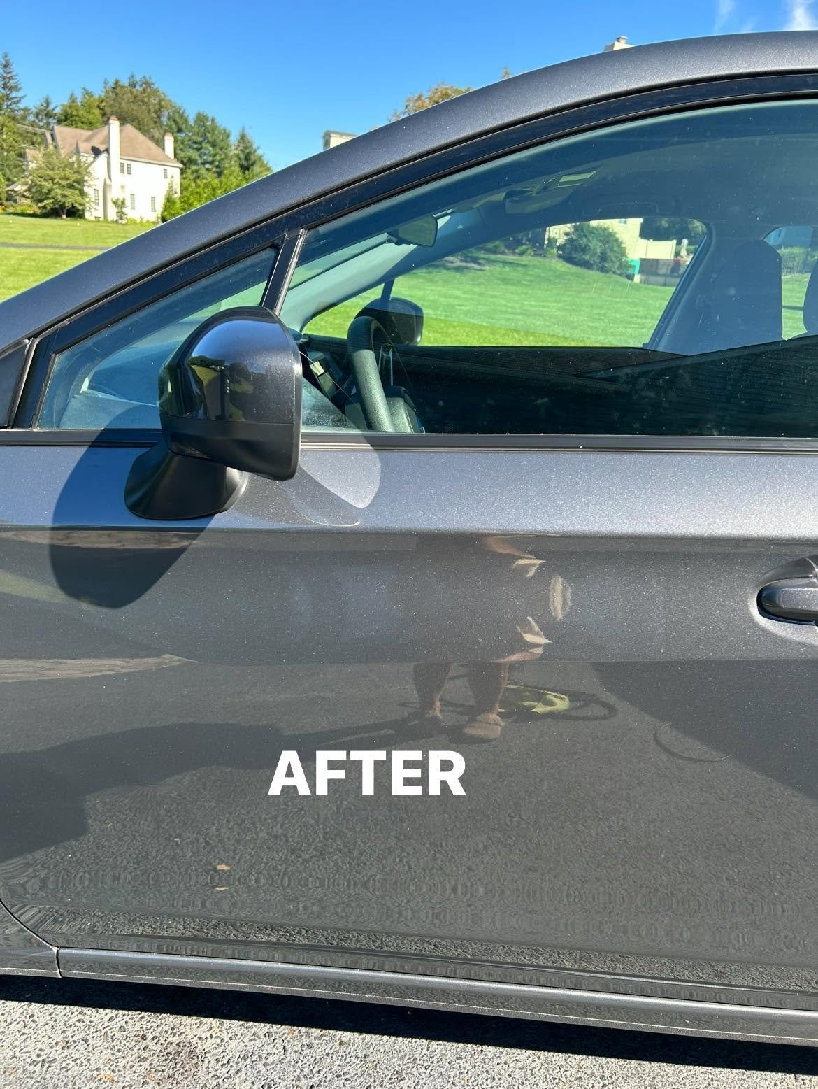 Side view of a gray car with a black mirror and open window after paint correction, showing a grassy field and house in the background