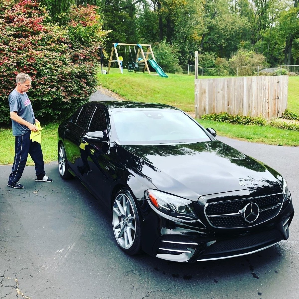 Detailer washing a shiny black Mercedes sedan in a residential driveway with a yard behind