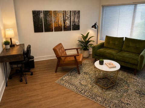 Modern living room with olive green sofa, mid-century wooden chair, white coffee table, desk with lamp, and forest wall art