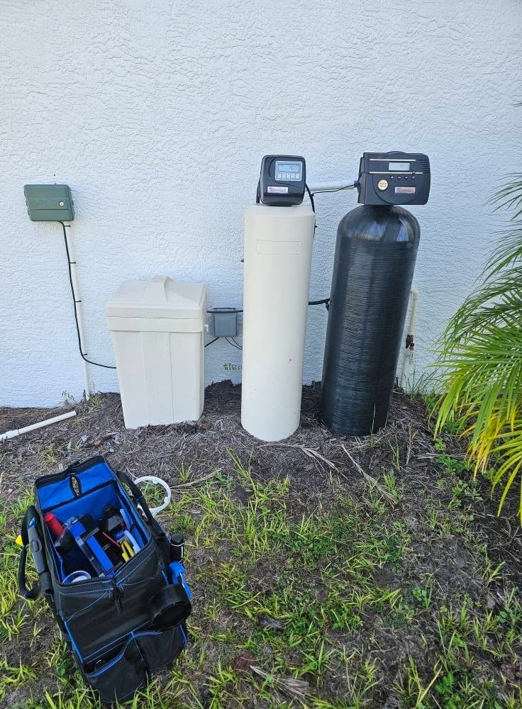 Water softener system with three tanks and digital controls mounted against white wall, with tool bag in foreground