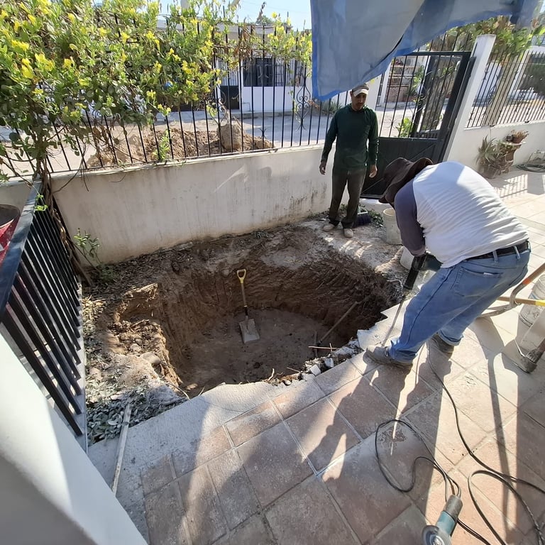 Two workers inspect a large rectangular excavation pit in a residential courtyard with iron gates and vegetation visible in the background