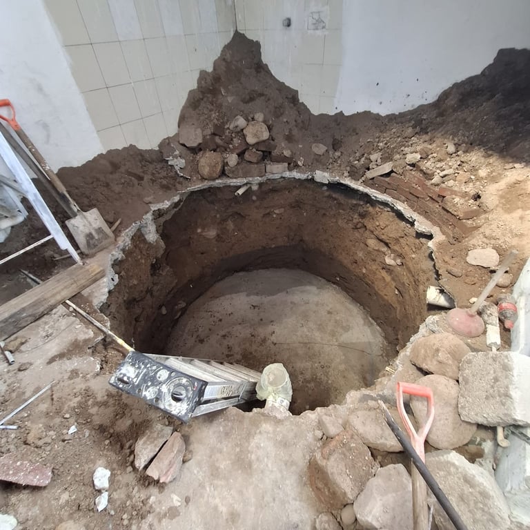 Large circular hole excavated in dirt floor with exposed concrete pipes and construction debris surrounding it