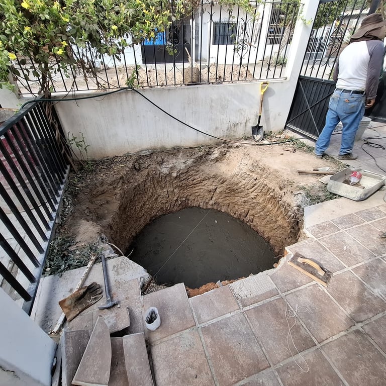 Large circular hole dug in residential driveway revealing underground sewer pipe during maintenance work