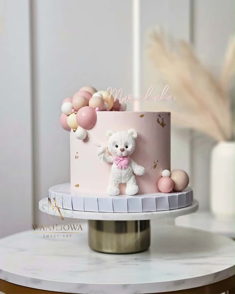 Pastel pink tiered cake decorated with a white teddy bear, pink balloons, and dried flowers on a marble surface