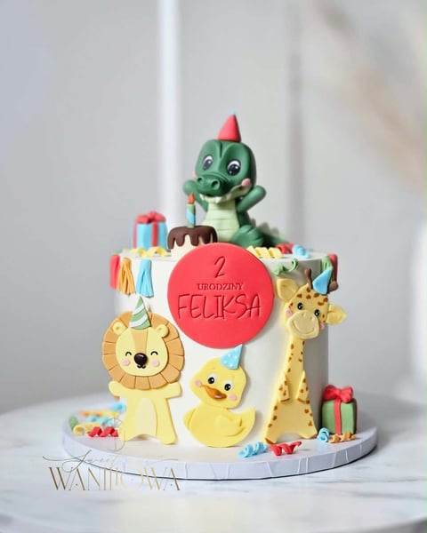Colorful childrens birthday cake decorated with cartoon animals including a dinosaur on top, lion, duck, and giraffe with a red circle featuring text