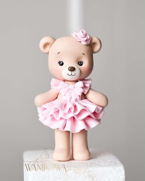 Cute bear figurine wearing a pink ruffled dress with a flower accessory on its head, standing on a white base