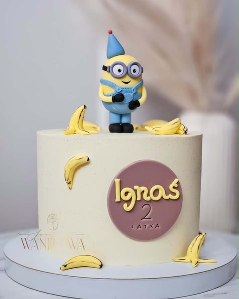 Birthday cake decorated with a Minion toy figurine wearing a blue party hat, gold bananas, and Ignos 2 Latra text on cream-colored fondant