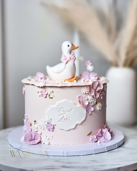 Pink tiered cake decorated with fondant flowers and a white duck figurine wearing a pink bow on top