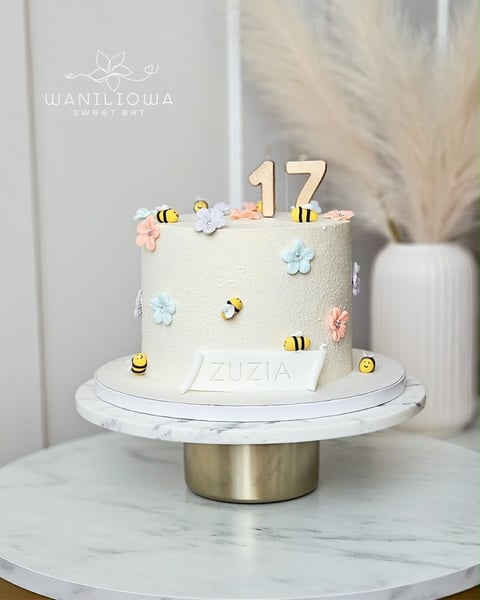 Cream-colored fondant cake decorated with bee and flower toppers, topped with gold number 17, on marble cake stand