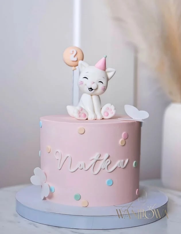 Pink birthday cake with white cat figurine wearing party hat holding number 2 balloon, decorated with clouds and confetti