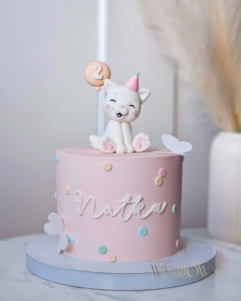 Pink birthday cake with a white cat topper holding a number 2 balloon, decorated with colorful polka dots and flowers