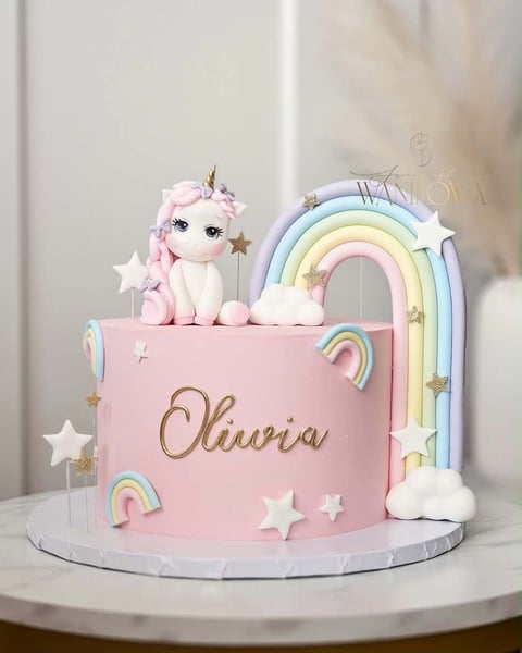 Pink unicorn birthday cake decorated with rainbow, clouds, stars, and the name Olivia written in gold script