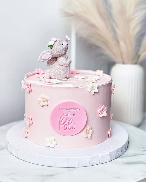 Pink birthday cake with white elephant figurine on top, decorated with small flowers and a personalized round plaque
