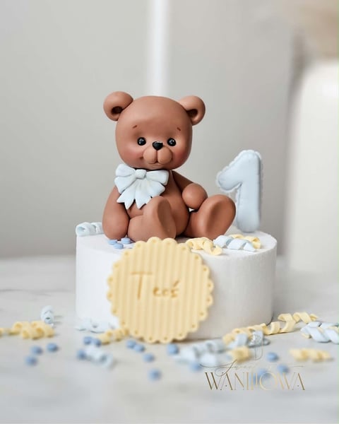 Brown teddy bear with white bow on white first birthday cake decorated with cream and blue pearl accents