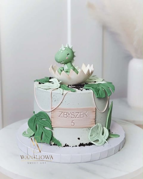 Green dinosaur hatchling cake with fondant details, wooden band, and tropical leaf decorations on white base