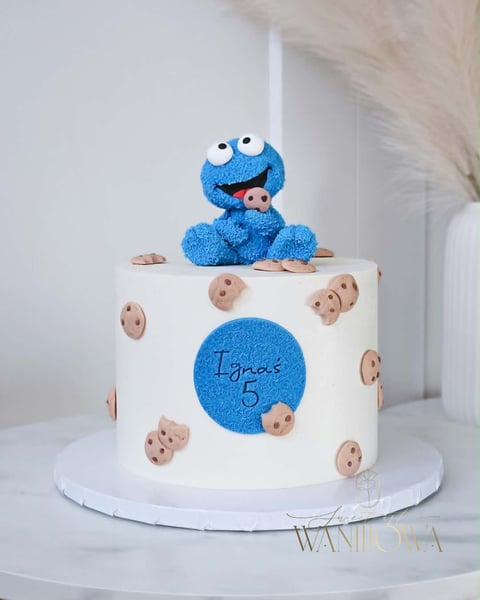 Blue Cookie Monster cake topper with white frosted cake decorated with cookie designs and blue circle, on white cake stand