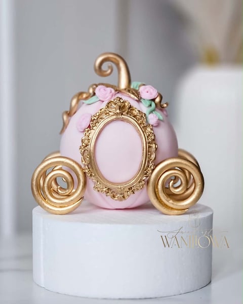 Decorative pumpkin carriage cake topper with pink body, gold accents, ornate oval frame, and floral details on white base