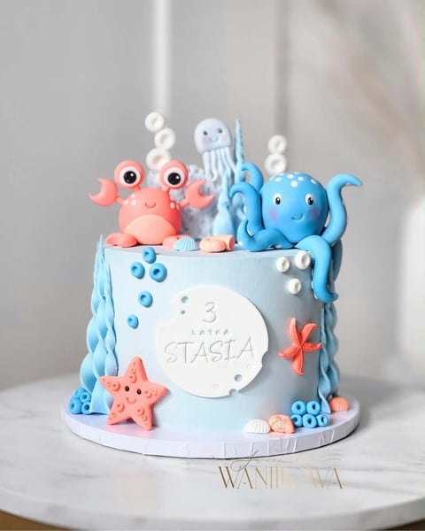 A light blue birthday cake decorated with sea creature toppers including a coral crab, blue jellyfish, and blue octopus, adorned with pearls and starfish