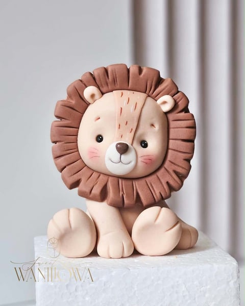 Cute fondant lion cake topper with brown mane, cream-colored face and body, sitting on a white cake