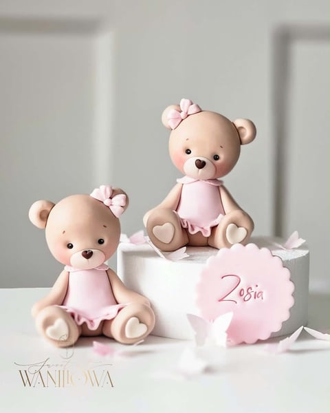 Two decorative teddy bears in pink dresses sitting on a white cake decorated with a pink tag reading Zosia
