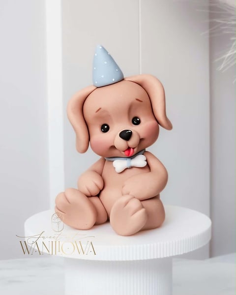 Cute fondant puppy cake topper with pink body, blue party hat, white bow tie, and tongue sticking out on white base