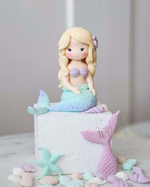 Fondant mermaid cake topper figurine with blonde hair sitting on a white tiered cake decorated with pastel shells and starfish