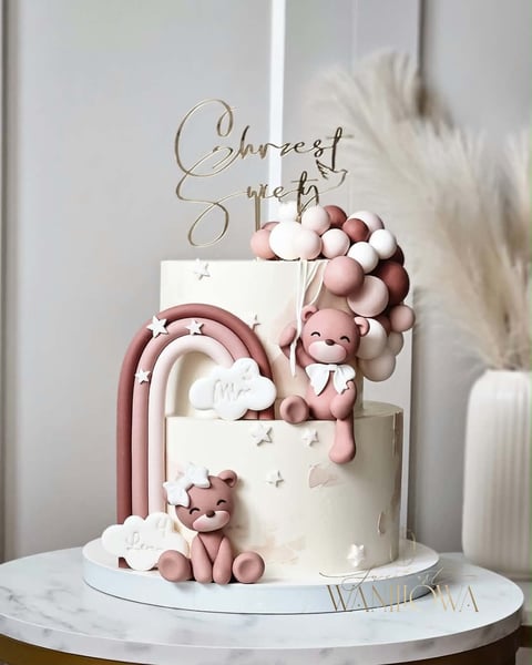 Elegant baby shower cake with pink and white tiers, teddy bear figurines, rainbow arch, and Sweetest Arrival cake topper on marble stand