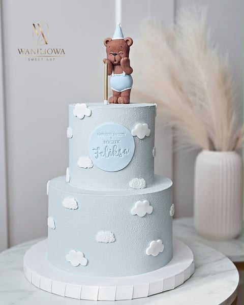 Two-tiered light blue birthday cake decorated with white clouds and topped with a brown teddy bear figurine wearing a party hat