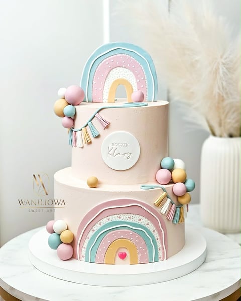 Two-tiered pastel cake with rainbow decorations, colorful fondant balls, and tassels on white stand