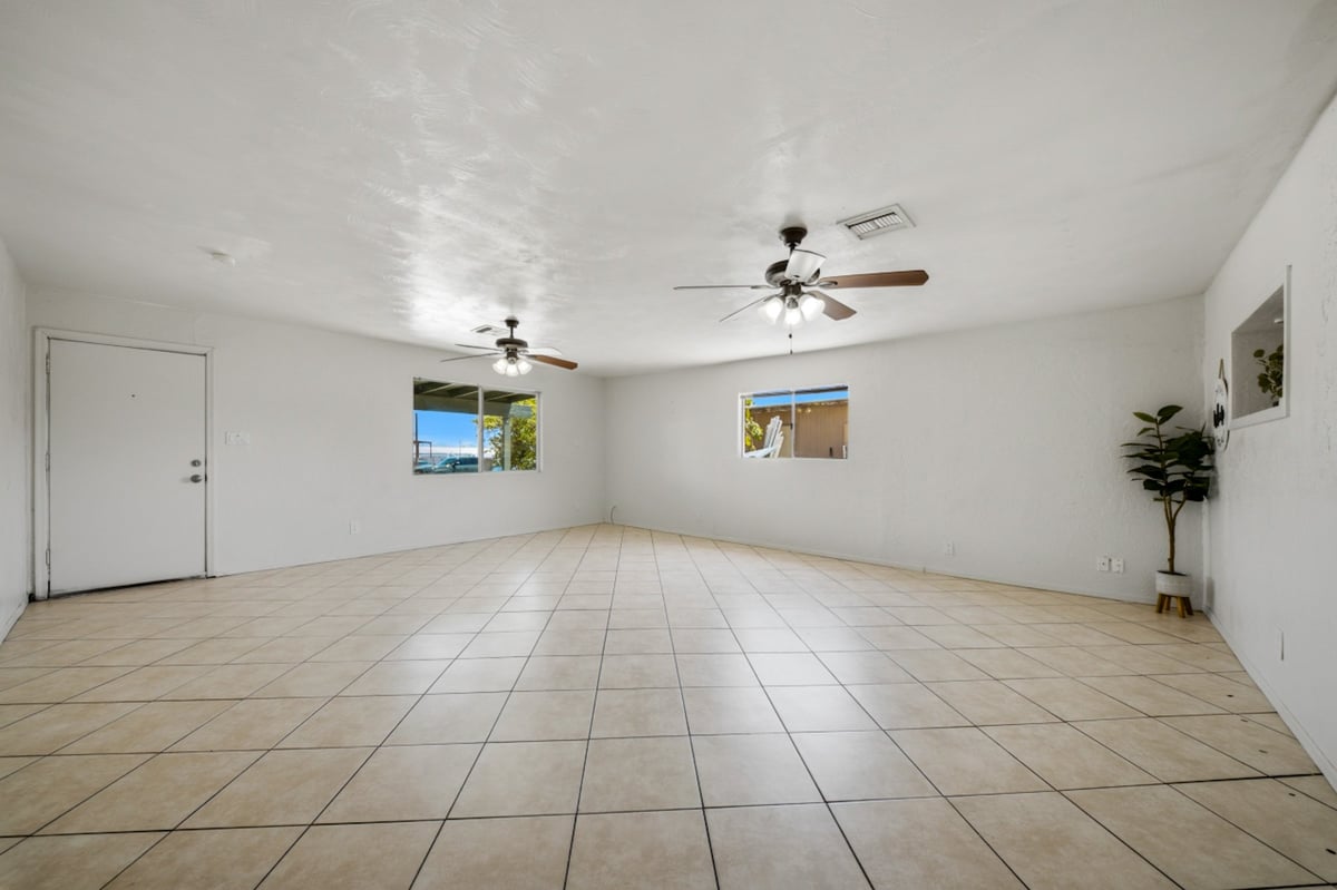 Empty living room with tile flooring, white walls, ceiling fans, and windows allowing natural light