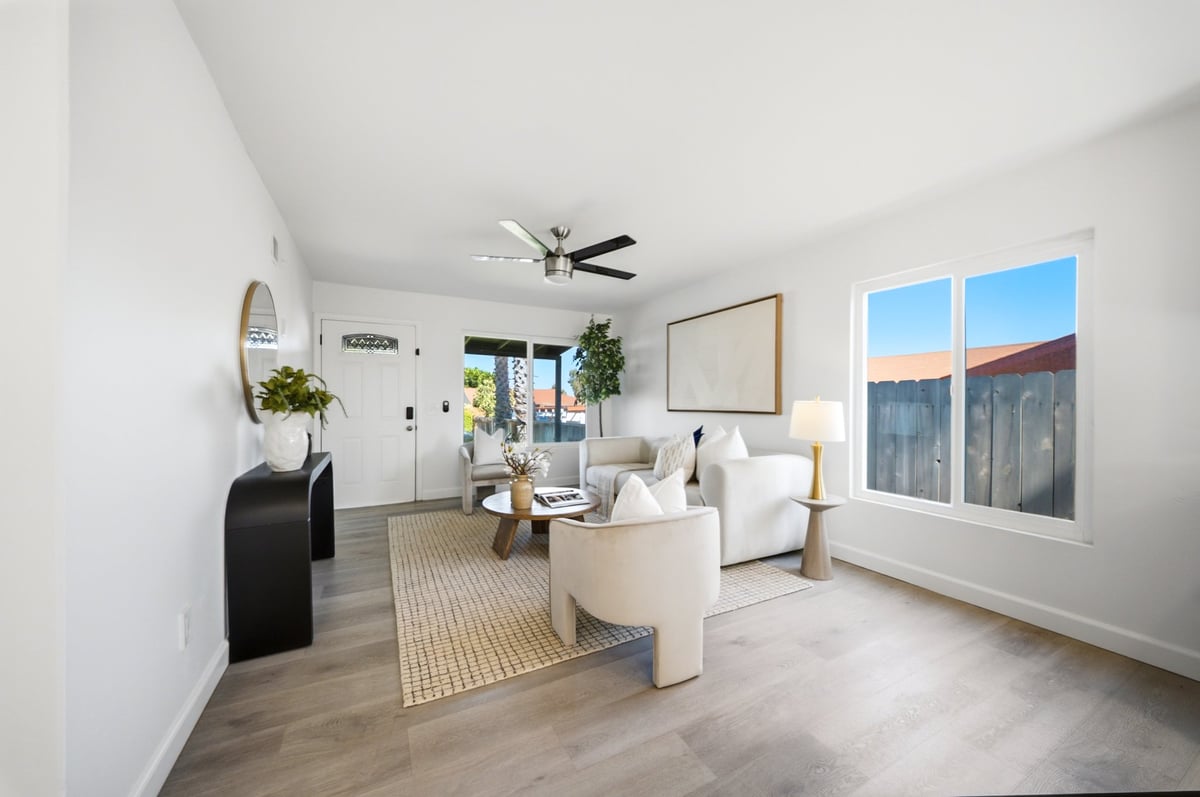 Modern living room with cream furniture, wood flooring, ceiling fan, and large windows overlooking a city skyline at sunset