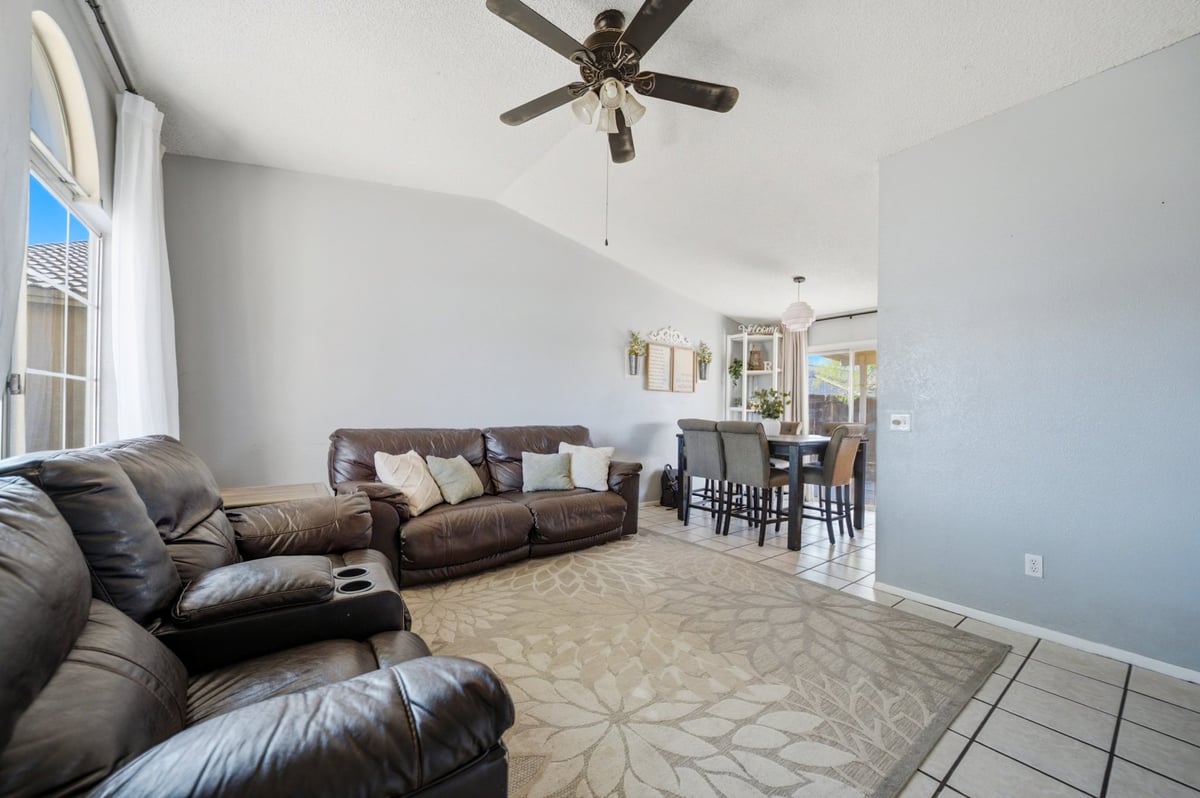 Modern open-plan living room with dark leather sectional sofa, ceiling fan, dining area visible in background, and tile flooring
